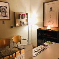 Photo shows the Red Door's syringe services office. Two pieces of Indigenous artwork hang on the walls next to a display case with brochures. The room is lit by a salt rock lamp and the soft yellow glow of a standing lamp. A desk is in the bottom right corner with two chairs in the bottom left. 